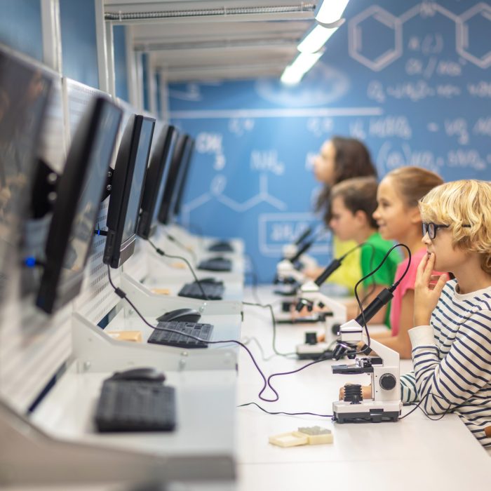 Information. Boy with glasses sideways to camera thinking looking at charging board and classmates in chemistry class